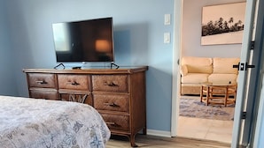 A bedroom with a bed, a dresser, a television, and a view into a living area with a sofa and a ceiling fan. - Beautifully renovated lakefront condo! Executive Rental, Monthly Rental (Lake Suzy)