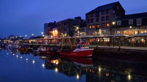 A waterfront with boats docked, a stone building with multiple windows, and a row of outdoor seating areas with umbrellas.