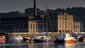 A marina with boats docked in front of a large, historic building with arched windows and a prominent chimney.