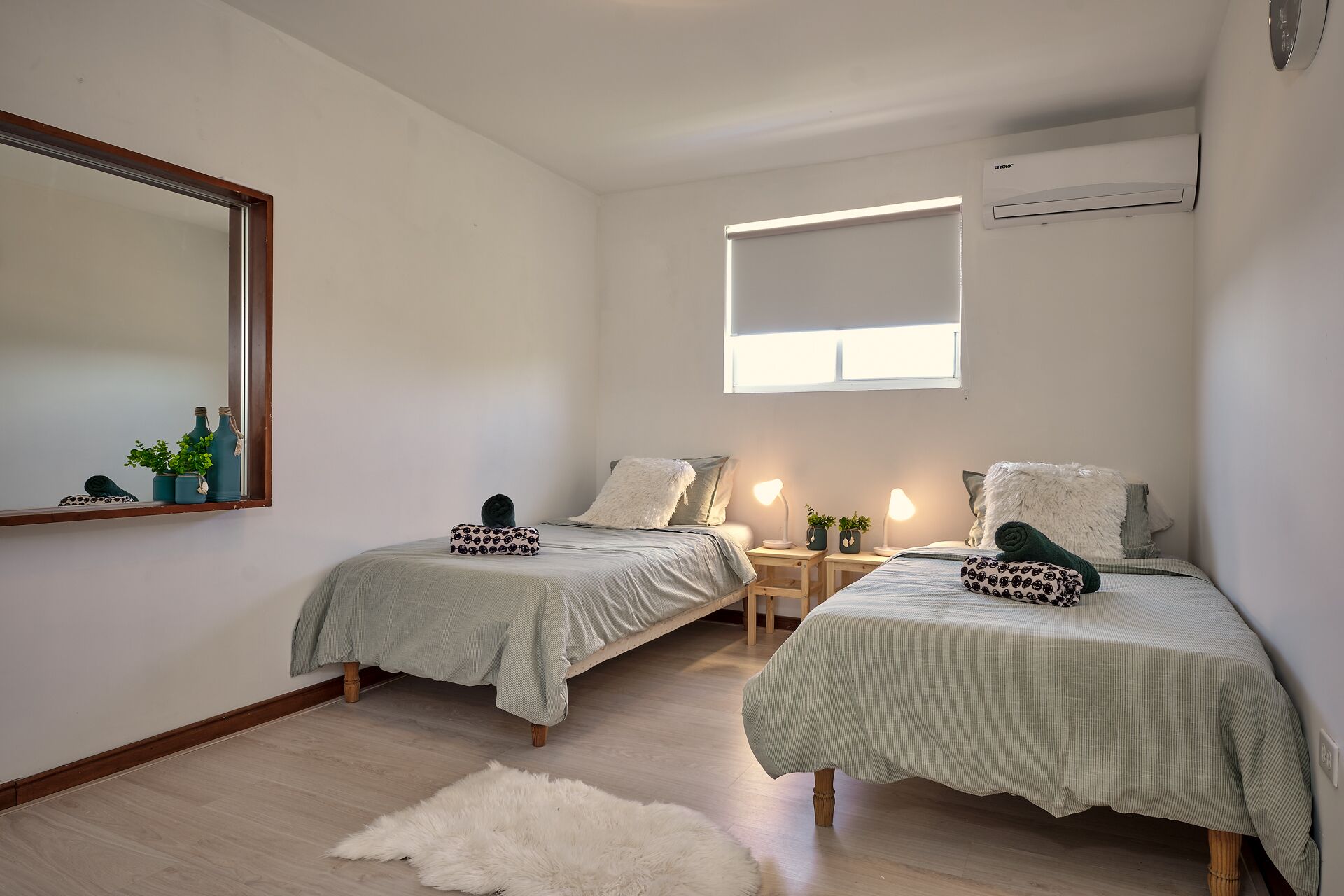A bedroom with two single beds, a large mirror, an air conditioning unit, and a small window.