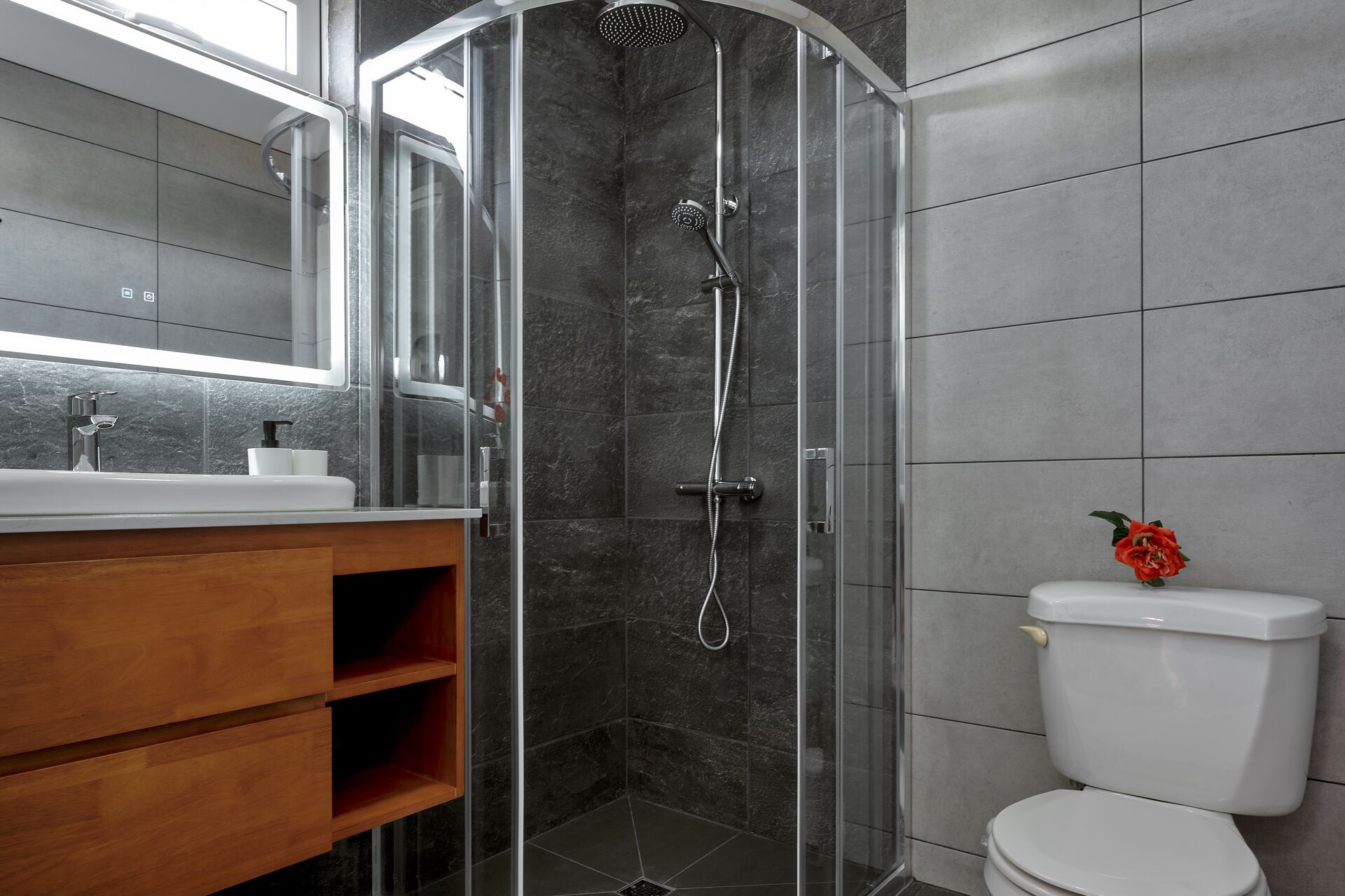 A modern bathroom with a glass-enclosed shower, a wooden vanity, and a toilet with a red flower on the tank.