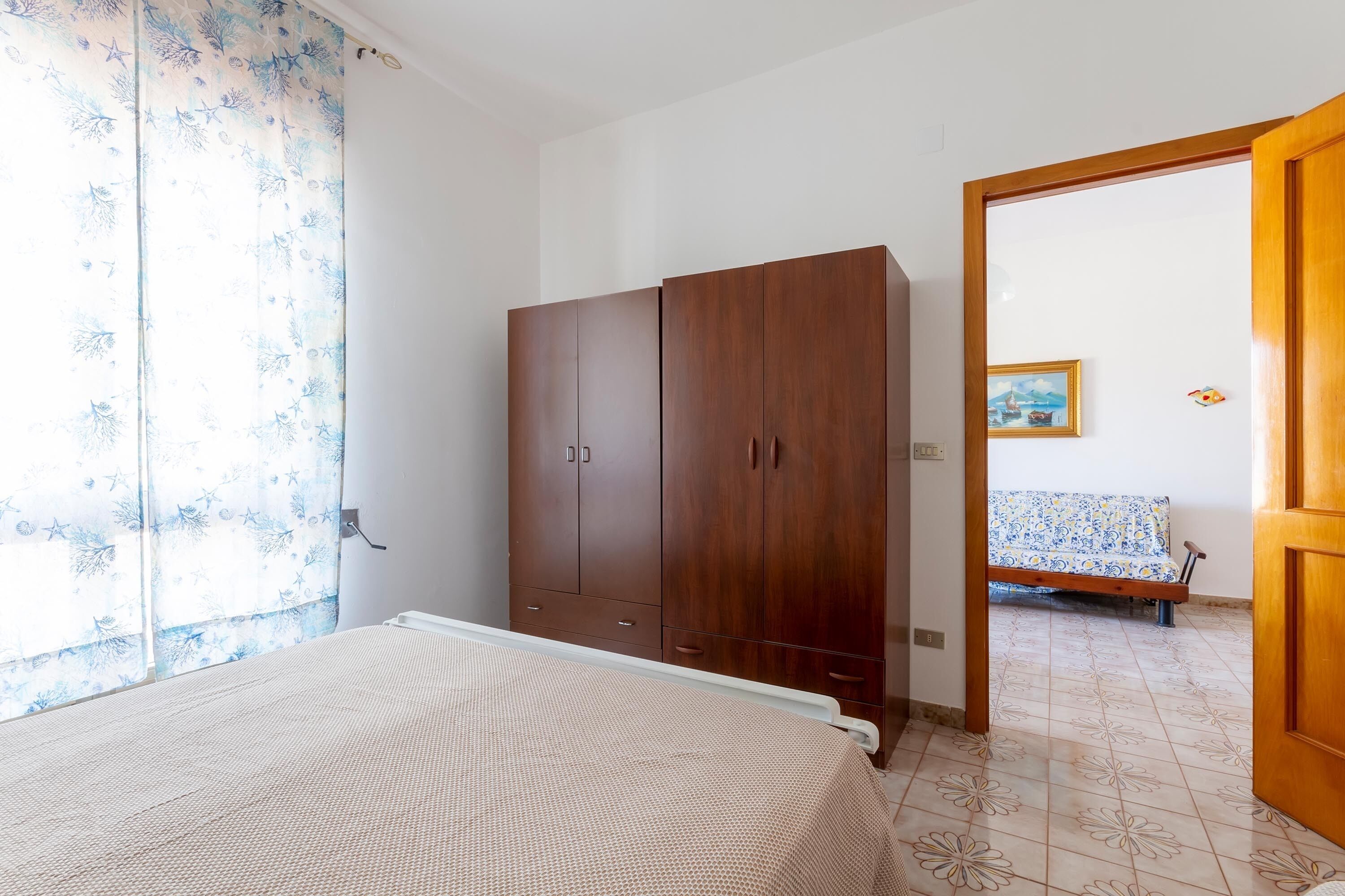 Quarto com cama, guarda-roupa de madeira e vista para um sofá no quarto vizinho.