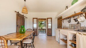 A rustic kitchen with a wooden table and chairs, a hanging light fixture, and shelves with kitchenware.