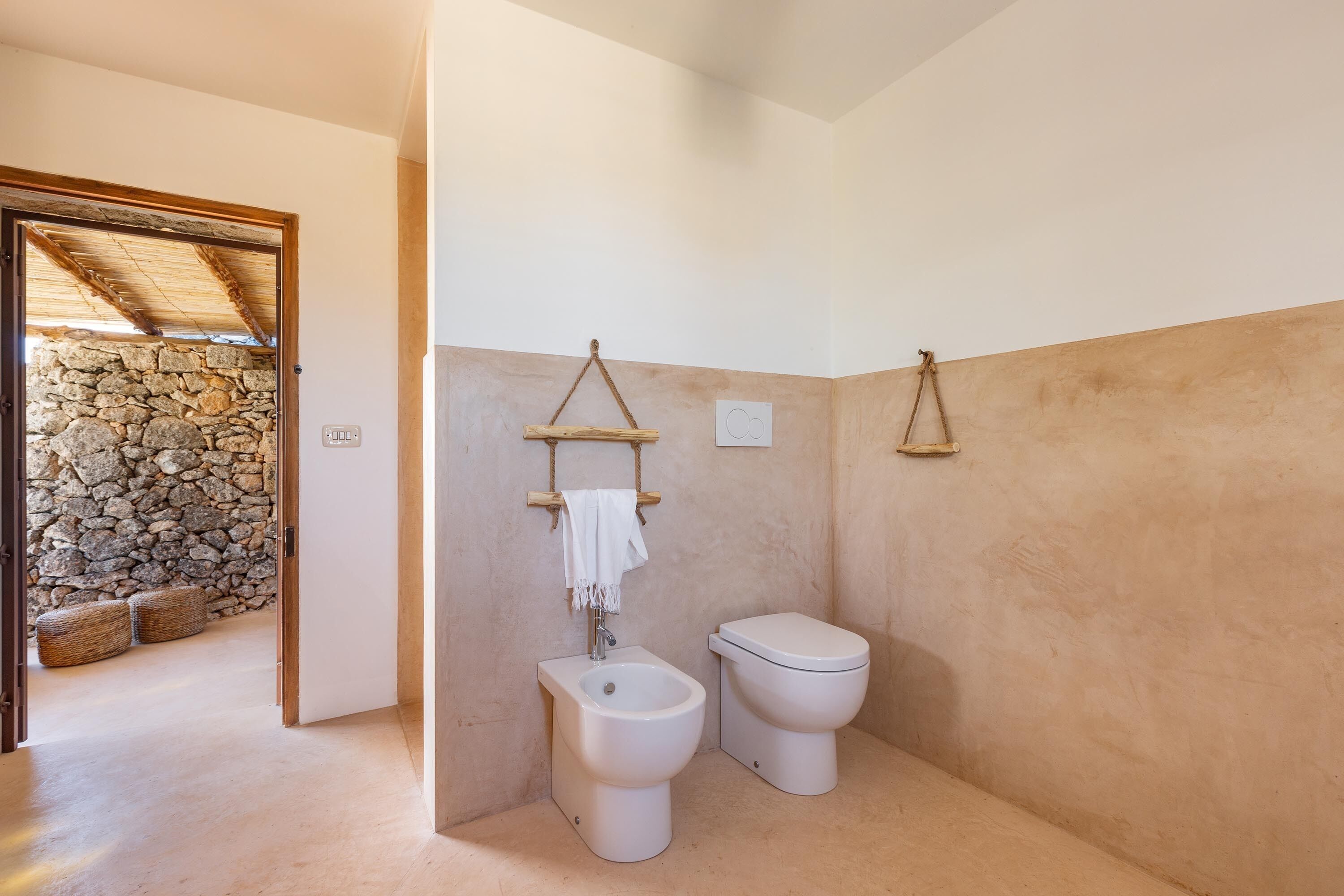 A bathroom with a stone wall, a wooden door, a bidet, a toilet, and a towel rack.