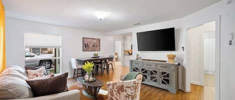 A living room with a sofa, a coffee table, and a patterned armchair. There is a dining area with a table and chairs, and a kitchen area with a cabinet and a flat-screen TV.