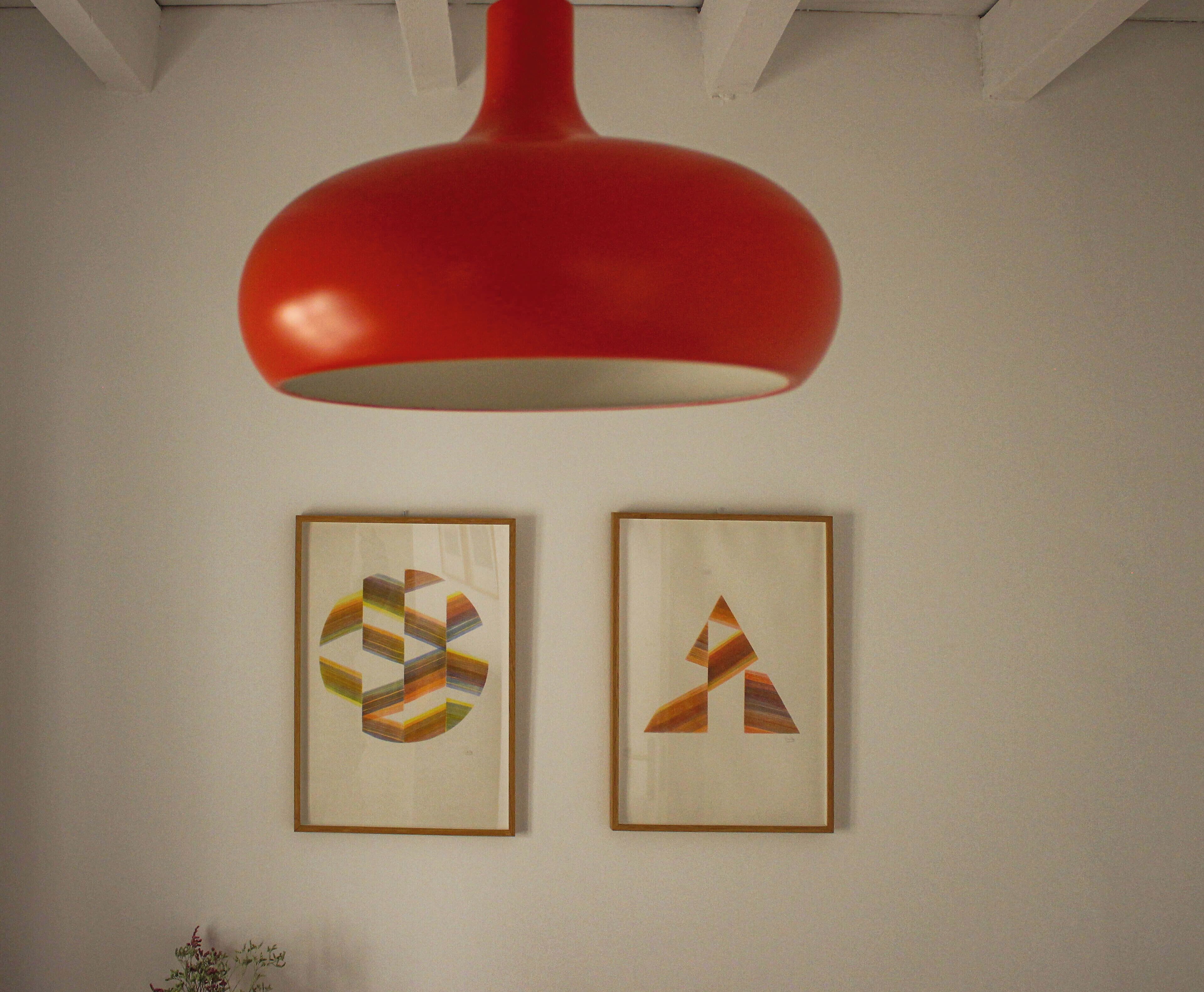 Dos obras de arte abstracto enmarcadas sobre una pared blanca, bajo una lámpara colgante roja.