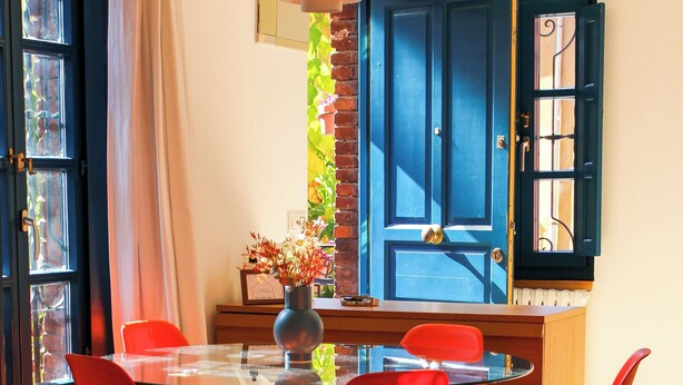 Un coin repas avec une table en verre, des chaises rouges, un vase avec des fleurs et une porte bleue avec des volets.