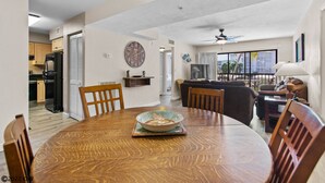 Un coin repas avec une table ronde en bois, des chaises et un bol décoratif. On aperçoit, au fond, une cuisine avec des placards et un réfrigérateur.