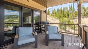 A wooden deck with two chairs and a grill, overlooking a forested area. - Luxury Ski In/Out ᨒ Beaverhead Condo | Hot Tub | 10 Min Walk to Resort (Big Sky)