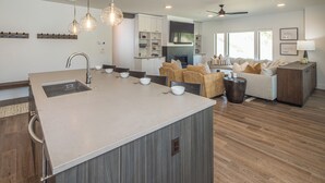 A modern kitchen with a central island, dining area with chairs, a sofa, and a TV wall-mounted above a fireplace. - Luxury Ski In/Out ᨒ Beaverhead Condo | Hot Tub | 10 Min Walk to Resort (Big Sky)