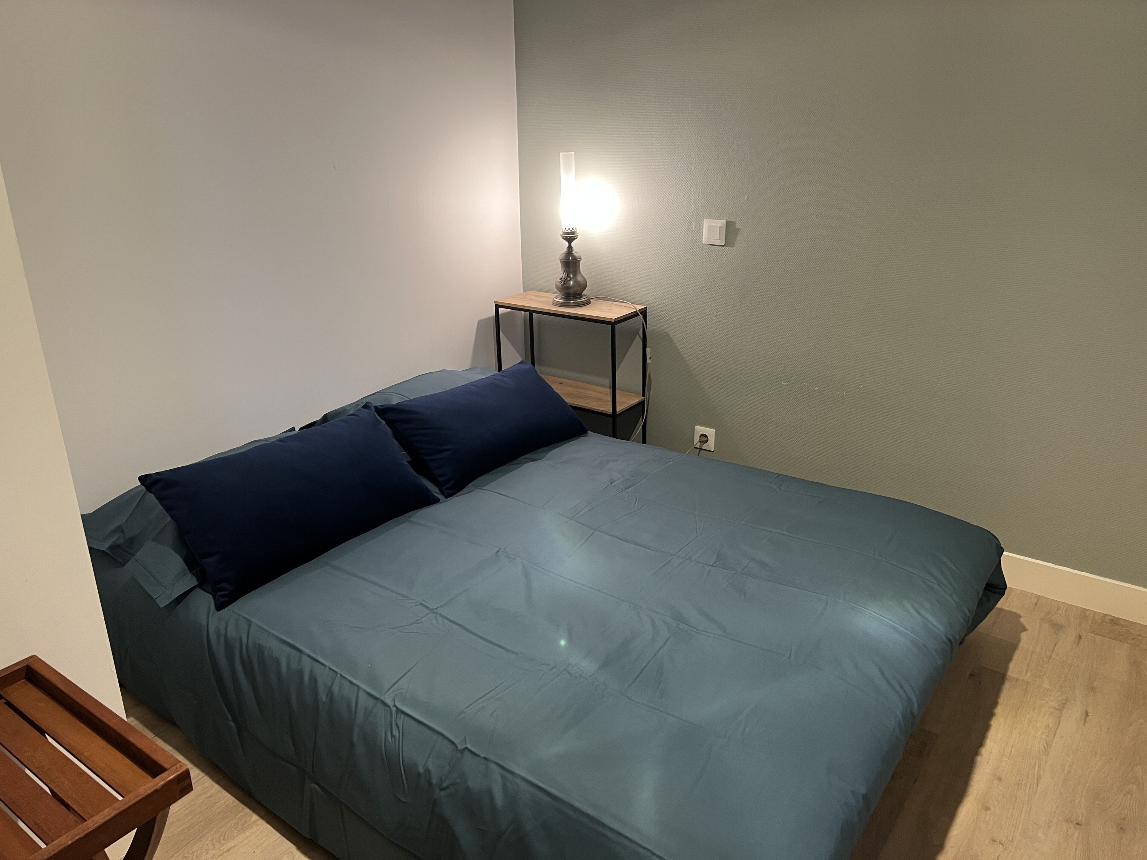 A single bed with a blue mattress and matching pillows, a small wooden nightstand with a lamp, and a power outlet on the wall.