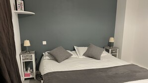 Ein ordentlich gemachtes Bett mit einer grauen Decke, zwei grauen Kissen und weißen Laken. Es gibt zwei Nachttische mit Lampen und ein kleines Regal über dem Bett.