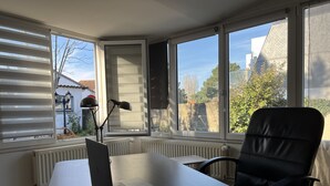 Ein Zimmer mit Schreibtisch, Laptop, Stuhl und großen Fenstern mit Jalousien.