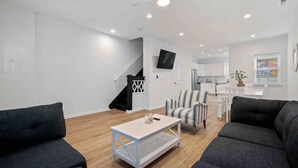 Un salon moderne avec un canapé, une table basse et un fauteuil à rayures. Un escalier avec une rampe noire mène à un espace cuisine doté de meubles blancs et d’un téléviseur fixé au mur.