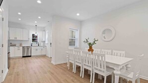 Une cuisine moderne avec des meubles blancs et des appareils en acier inoxydable, un coin repas avec des chaises et une table blanches, et une petite fenêtre avec des stores.