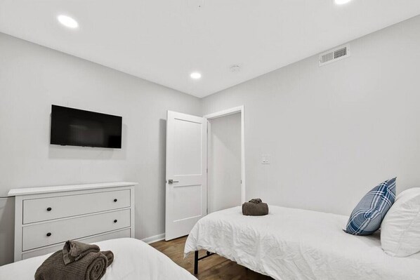 A bedroom with two beds, a TV mounted on the wall, a white dresser, and a door leading to another room.
