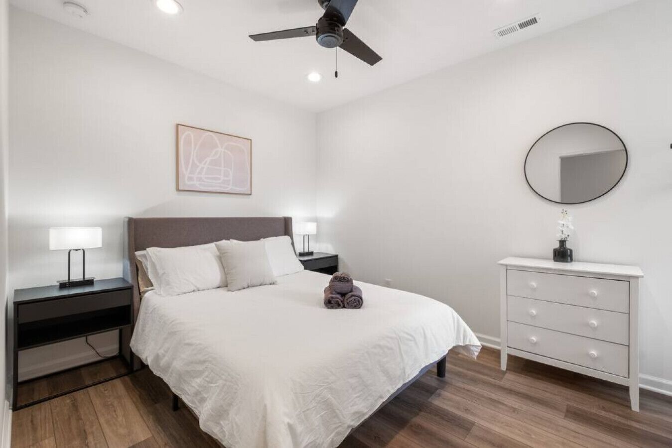 Un dormitorio con cama, mesitas de noche, cómoda, ventilador de techo y espejo en la pared.