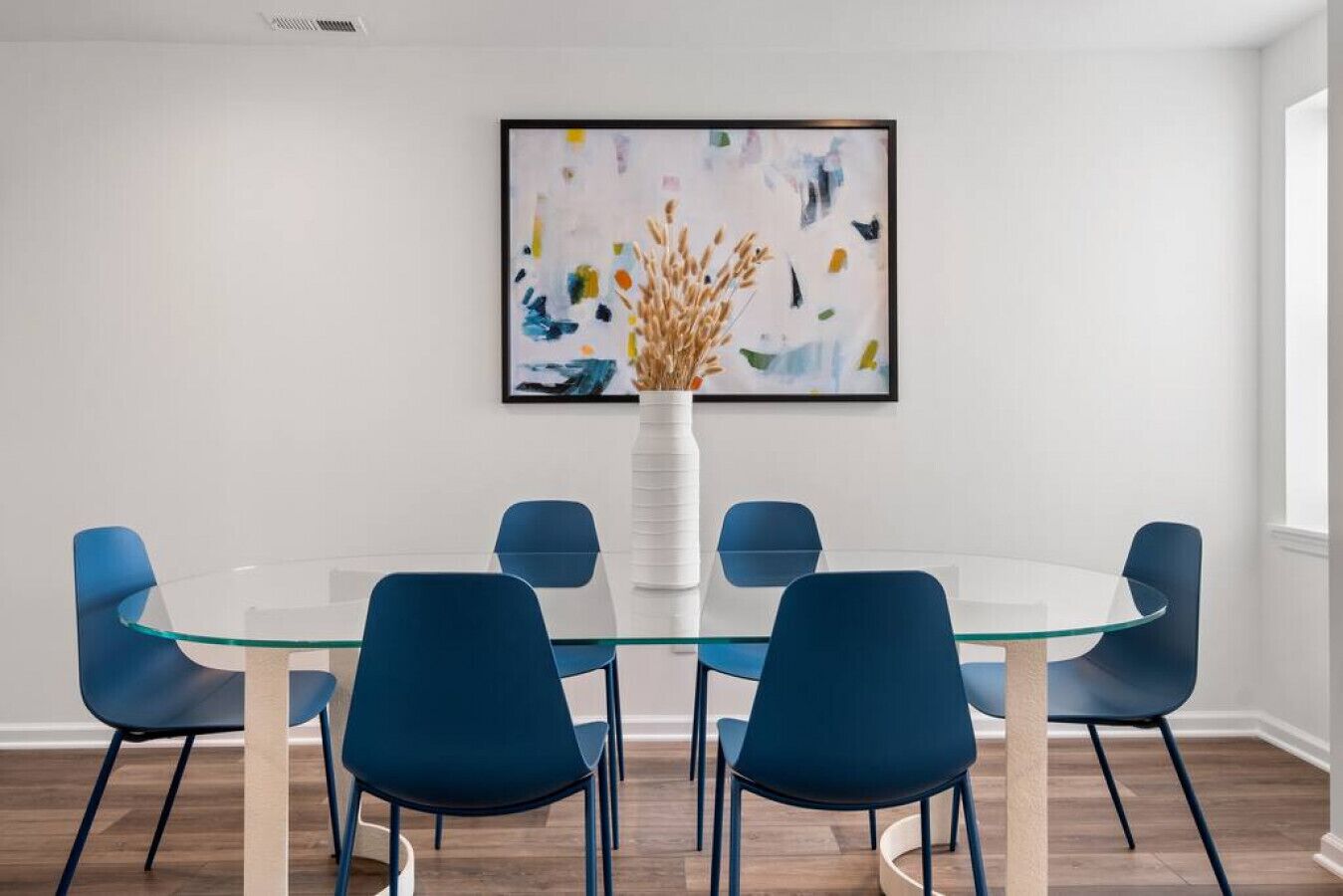 Un comedor moderno con una mesa de vidrio rodeada por seis sillas azules, un jarrón blanco con plantas secas y un cuadro abstracto y colorido en la pared.