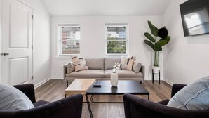 A modern living room with a beige sofa, a coffee table, and two armchairs. There are two windows with blinds, a large plant, and a flat-screen TV mounted on the wall.