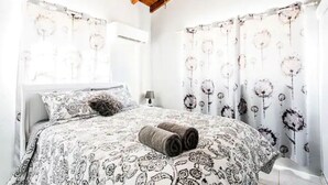 Ein Schlafzimmer mit Bett, gemusterten Betttextilien, gerollten Handtüchern, einem Nachttisch und blumigen Vorhängen.