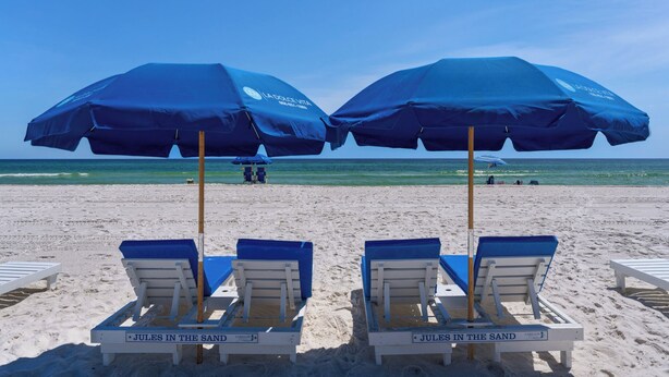 En strand med to blå parasoller, liggestole og et skilt med teksten "Jules in the Sand".