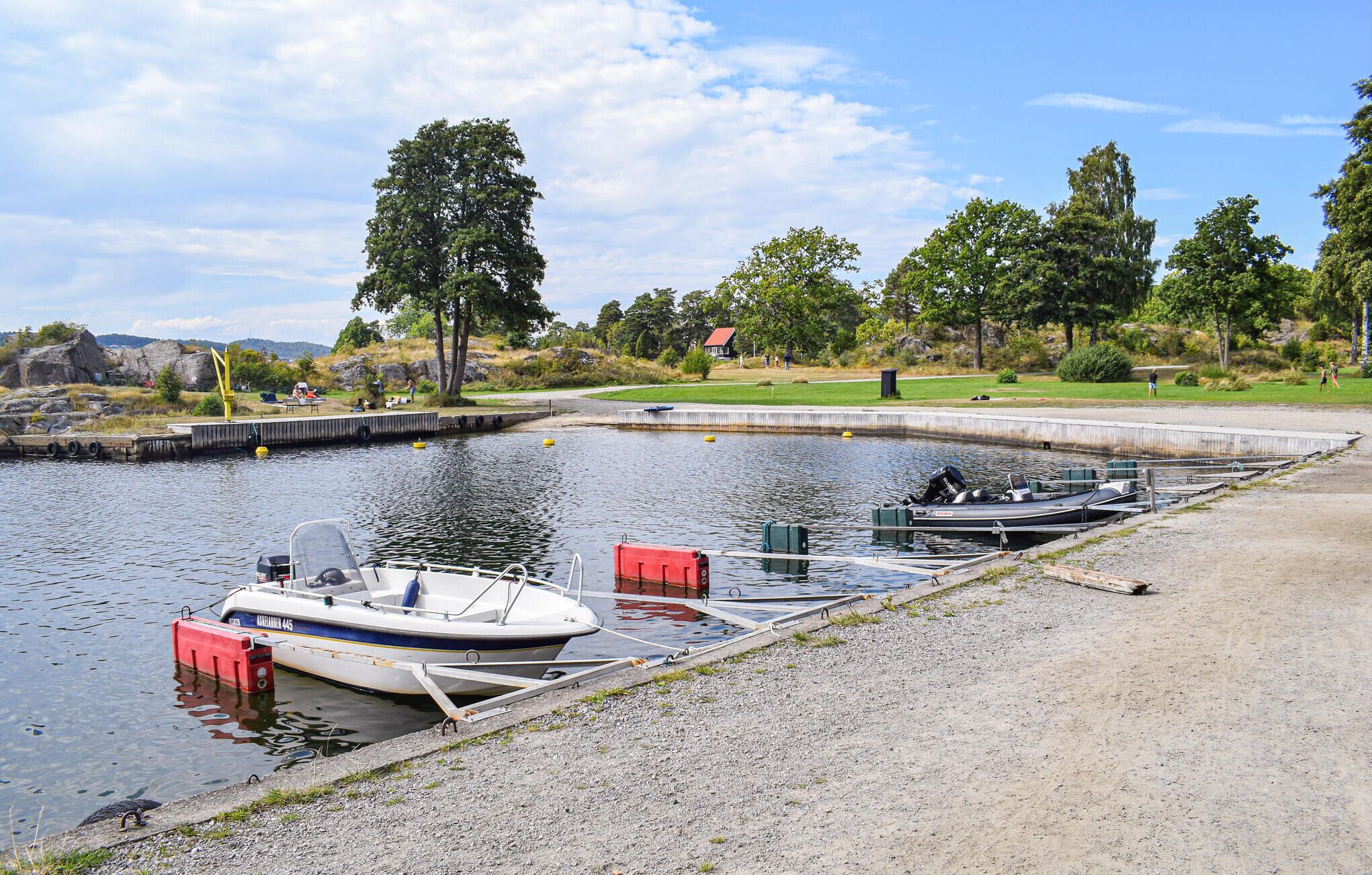 A calm lake with boats docked at a concrete pier, surrounded by trees and a grassy area with a small building in the background.