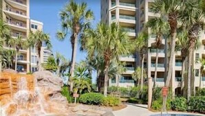 A hotel with multiple floors, palm trees, and a pool area with a waterfall feature. - Sandestin, Gulf Front Condo, Golf Cart (Miramar Beach)