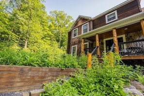 A two-story wooden house with a porch, surrounded by lush greenery and trees.