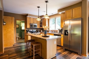 A modern kitchen with wooden cabinets, stainless steel appliances, a central island with stools, pendant lighting, and a view of the outdoors.