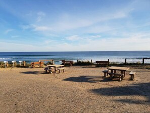 Ein Strand mit Holzliegen und Picknicktischen, ein Sandufer und das Meer im Hintergrund.