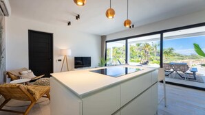 Une cuisine moderne dotée d’un grand îlot central, d’un coin repas avec des chaises, d’un téléviseur et offrant une vue sur l’extérieur.