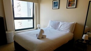 A neatly made bed with white linens and a rolled towel, a window with a view, a wall-mounted picture, and a small bedside table with a lamp. - No 2 - Modern 2 Bed Apartment, Close to Gla Airport & M8 (Paisley)