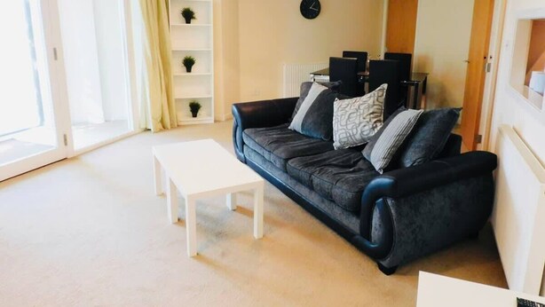 A living room with a black sofa, white coffee table, and a white shelving unit with plants. - No 2 - Modern 2 Bed Apartment, Close to Gla Airport & M8 (Paisley)