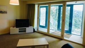 A room with a television on a white stand, a table, and large sliding glass doors leading to a balcony with a view of greenery. - No 2 - Modern 2 Bed Apartment, Close to Gla Airport & M8 (Paisley)