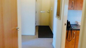 A hallway leading to a door, a small kitchen area with cabinets, and a black mat on the floor. - No 2 - Modern 2 Bed Apartment, Close to Gla Airport & M8 (Paisley)