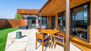 An outdoor patio with a wooden table and chairs, a trash can, and a view of a modern building with large windows. - Joy of Life @ Palisade Legends (Clifton)