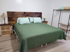 A neatly made bed with a green bedspread, two pillows, and a wooden headboard. There are two bedside tables with lamps and a small shelf unit with a hanging item.