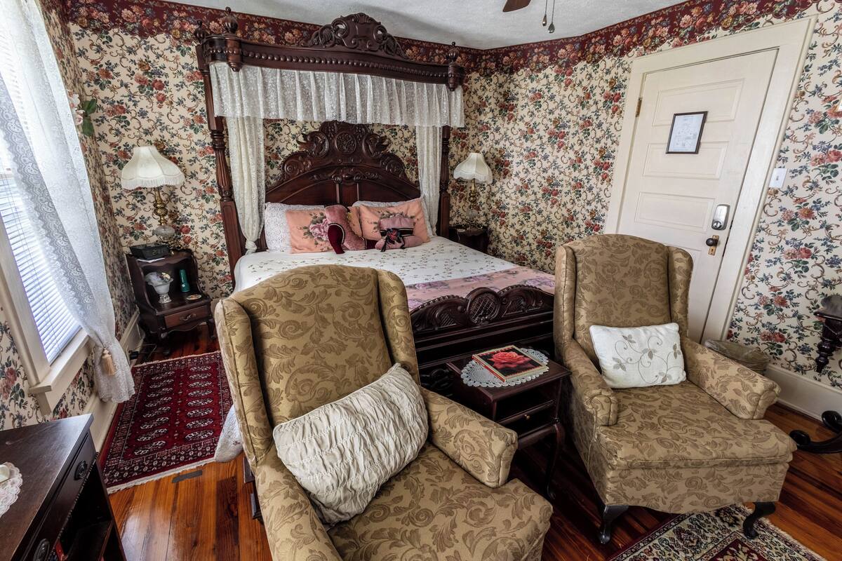 Une chambre à coucher avec un lit à baldaquin, un papier peint à motifs floraux, deux fauteuils, un plancher en bois et une porte ornée d’un cadre.