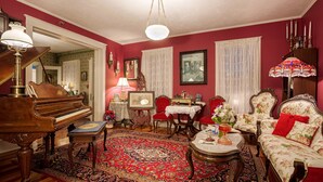 A traditional living room with a grand piano, red floral sofa, ornate table, and a patterned rug. - Alling House (Orange City)