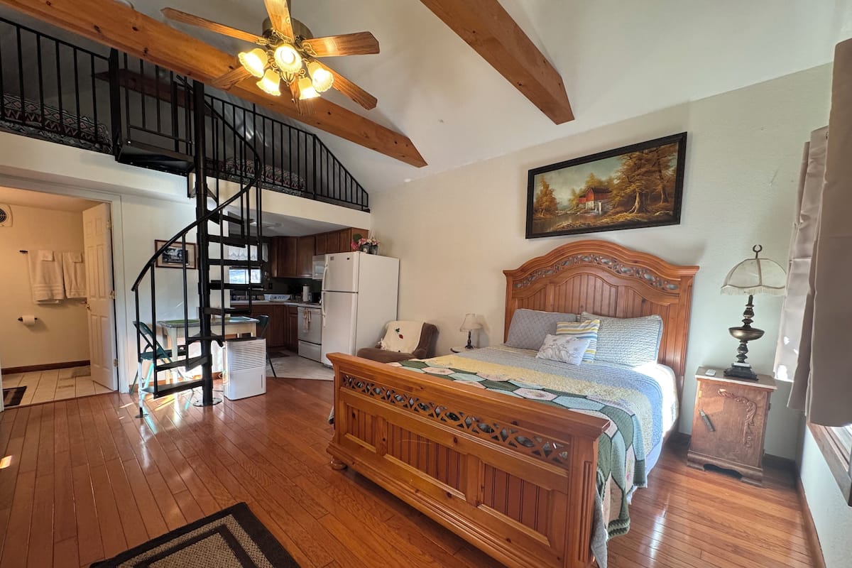 Une chambre avec un lit en bois, un ventilateur de plafond, un tableau au mur et un coin cuisine doté de meubles en bois et d’un réfrigérateur.