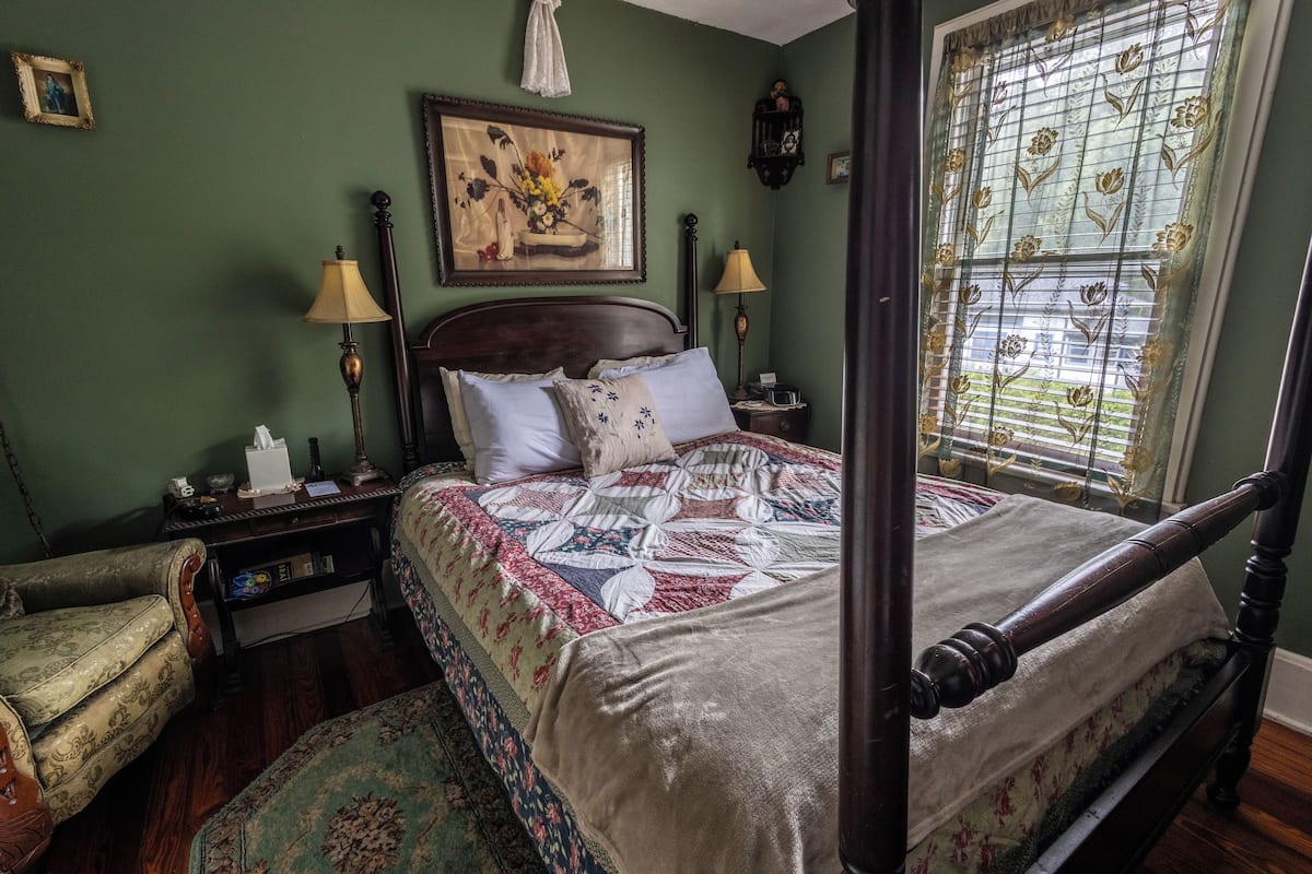 Un lit à baldaquin avec une courtepointe à motifs floraux, une table de chevet en bois avec une lampe, une chaise et un tableau accroché au mur.
