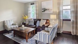 A living room with a brown leather sofa, striped armchairs, a wooden coffee table, and a large abstract painting on the wall. - Charming Carnegie Cottage 15 Minutes from Airport & Downtown (Carnegie)