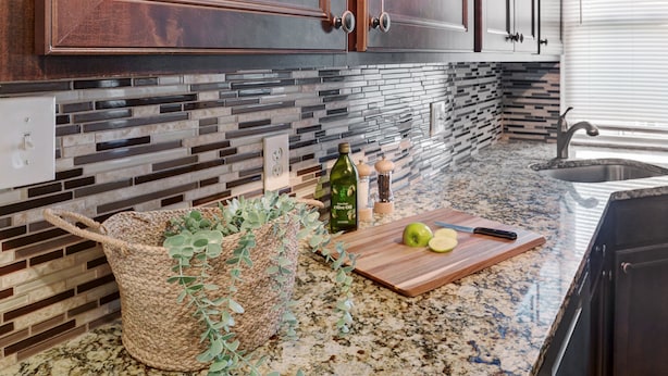 A kitchen with a granite countertop, a woven basket, a cutting board with apples, and a bottle of olive oil. - Charming Carnegie Cottage 15 Minutes from Airport & Downtown (Carnegie)