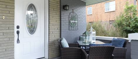 A covered porch with a white door, wicker furniture, a small table, and a welcome mat.