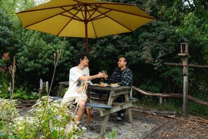 Two people dining at an outdoor wooden table with a yellow umbrella, surrounded by greenery.