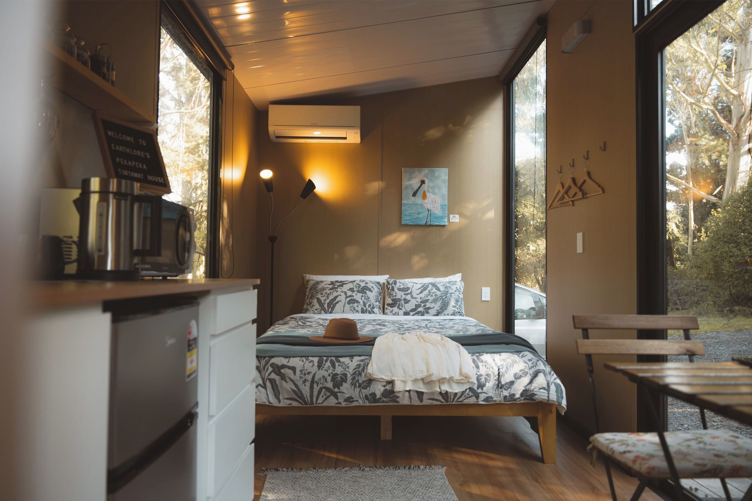 Un dormitorio con cama, escritorio con cafetera, un cuadro en la pared y vistas a árboles a través de la ventana.