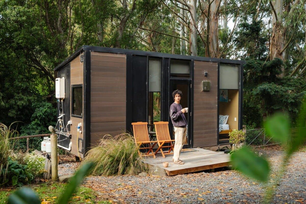 A modern cabin with a wooden exterior, a small porch with two chairs, and a person standing at the entrance.