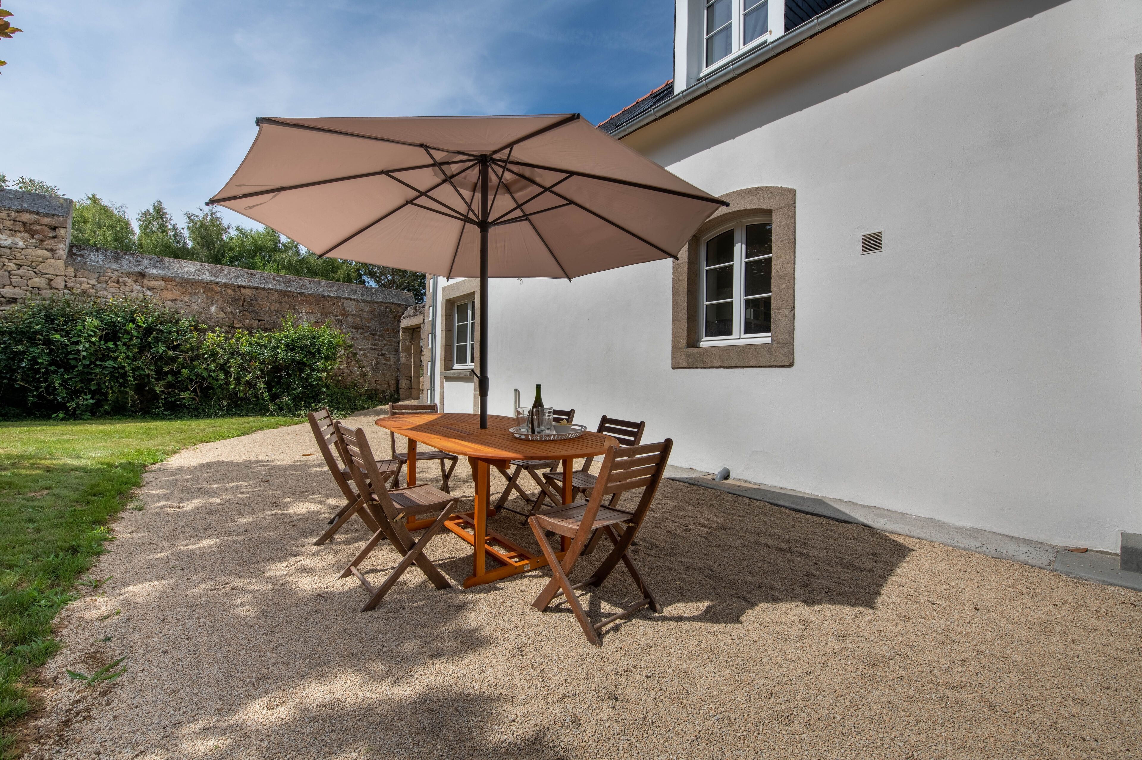 Une terrasse avec une table et des chaises en bois, un grand parasol et un mur de pierre en arrière-plan.