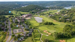 An aerial view of a golf course with residential areas, a large building complex, and a lake in the background.