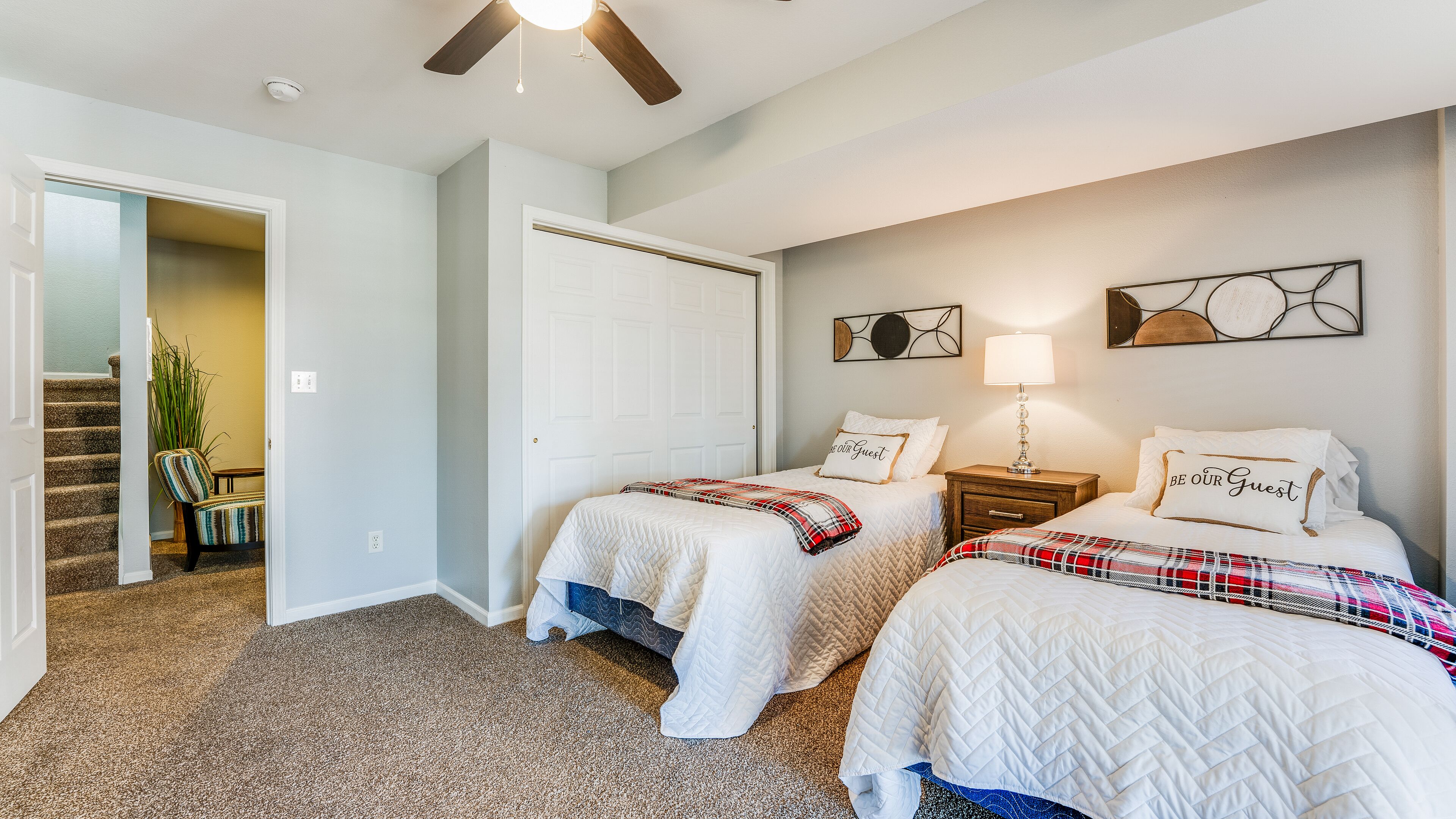 A bedroom with two beds, a ceiling fan, a nightstand with a lamp, and a door leading to a staircase.